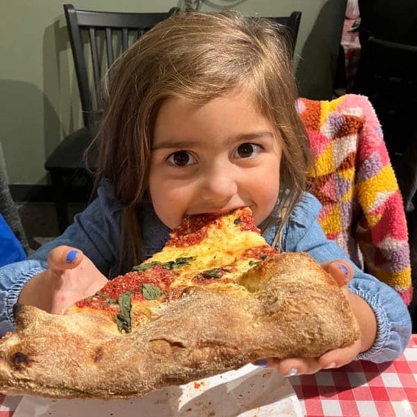 A young girl with brown hair takes a big bite out of a large slice of Casa Pizzeria's craft pizza.