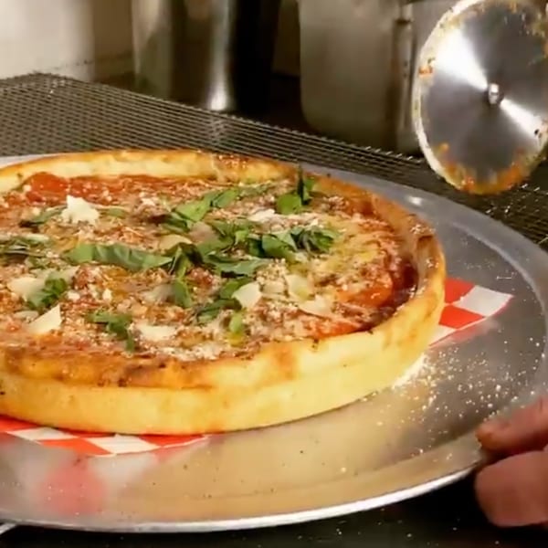 Deep-dish pizza with basil and cheese on a metal tray, being cut by a pizza wheel.