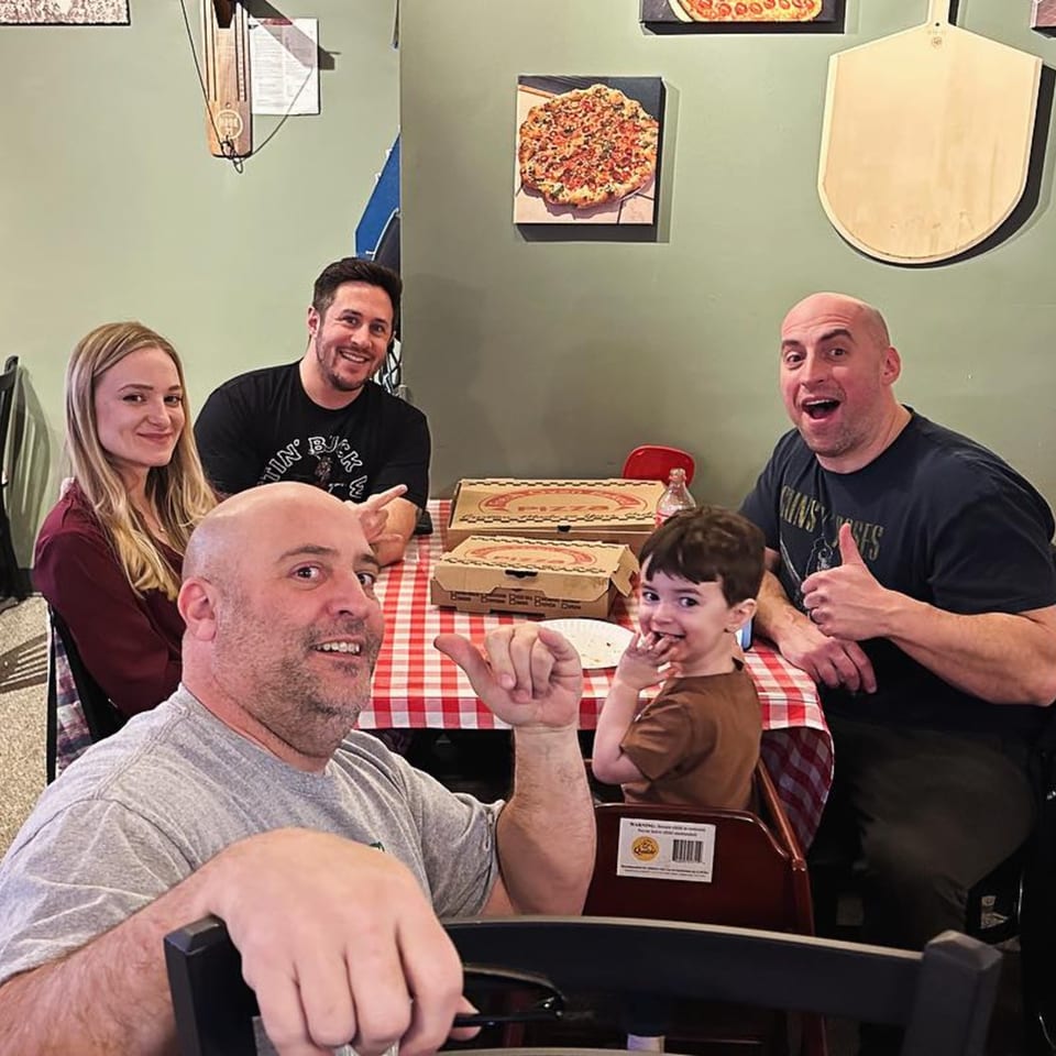 A family of five, including a young child, happily poses at a table with pizza boxes.