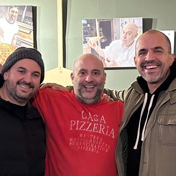 Three smiling men, one in a red Casa Pizzeria shirt, stand together in front of pizza-themed photos.