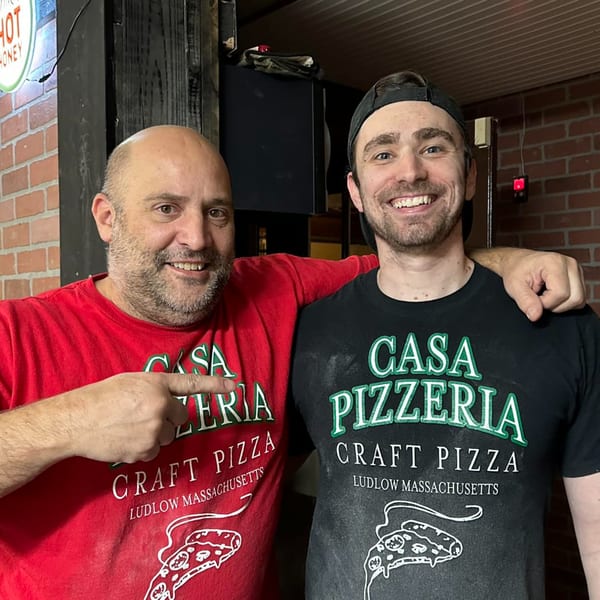 Two smiling men in Casa Pizzeria shirts, one pointing to his shirt, standing in front of a brick wall.